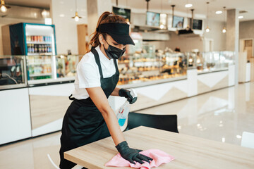 Beautiful woman working bakery or fast food restaurant. She is cleaning and disinfecting tables against Coronavirus pandemic disease. She is wearing protective face masks, gloves and face shield.