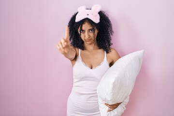 Hispanic woman with curly hair wearing sleep mask and pajama holding pillow pointing with finger up and angry expression, showing no gesture