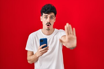 Hispanic man using smartphone over red background doing stop gesture with hands palms, angry and...
