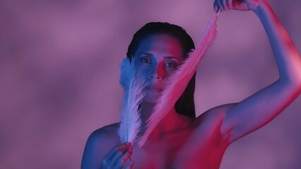 Close up shot of a seminaked young woman touching her face with two feathers in blue and pink color scheme and shadowed background.