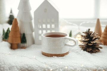 Obraz na płótnie Canvas Winter hygge. Stylish cup of tea with modern cute christmas houses, pine cone, wooden star and tree, golden lights on soft warm blanket on windowsill. Christmas cozy still life. Merry Christmas!
