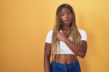 African american woman with braided hair standing over yellow background surprised pointing with finger to the side, open mouth amazed expression.
