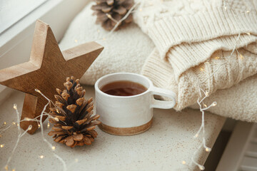 Fototapeta na wymiar Winter and autumn hygge. Stylish cup of tea with cozy knitted sweater, pine cone, wooden star and golden lights on windowsill in festive scandinavian room. Cozy Christmas. Happy Holidays!