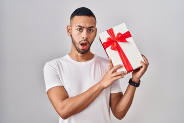 Young hispanic man holding presents in shock face, looking skeptical and sarcastic, surprised with...