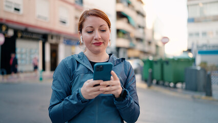 Young redhead woman wearing sportswear listening to music using smartphone at street