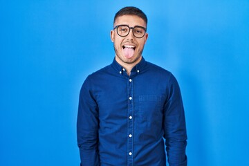 Young hispanic man wearing glasses over blue background sticking tongue out happy with funny expression. emotion concept.