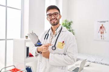 Young hispanic man doctor smiling confident holding clipboard at clinic