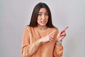 Young brunette woman standing over white background pointing aside worried and nervous with both...