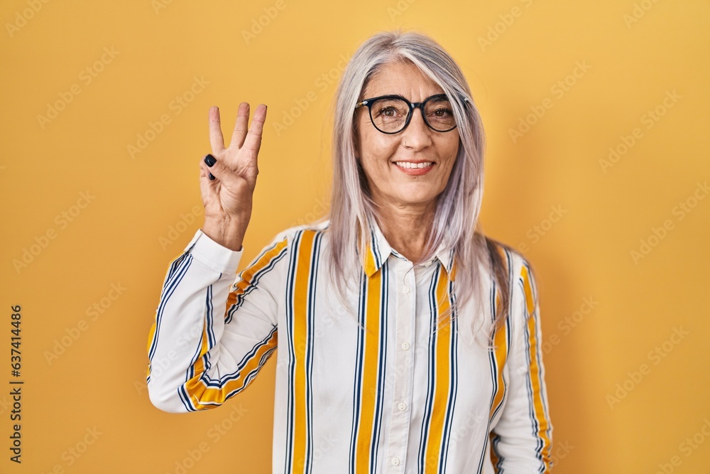 Wall mural middle age woman with grey hair standing over yellow background wearing glasses showing and pointing