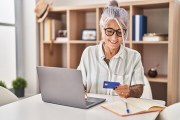 Middle age grey-haired woman using laptop and credit card sitting on table at home