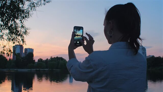 Woman takes photo of lake with mobile phone. Beautiful girl takes pictures and videos on her phone of a sunset on summer evening