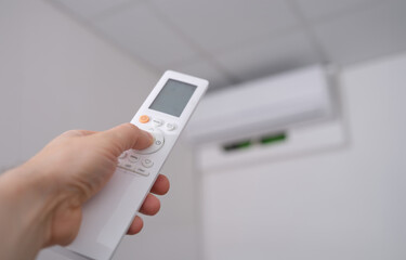 Woman hand holds remote control pointing at air conditioner in room. Adjusted to ambient temperature
