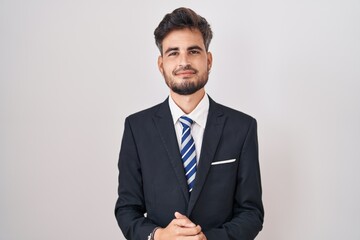 Young hispanic man with tattoos wearing business suit and tie with hands together and crossed fingers smiling relaxed and cheerful. success and optimistic