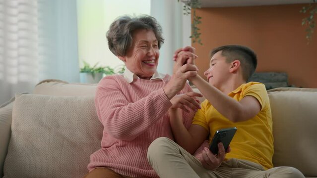 Happy funny caucasian grandmother and grandson playing mobile phone together at home couch give high five hands grandma with kid boy celebrating victory in online game on smartphone teamwork success