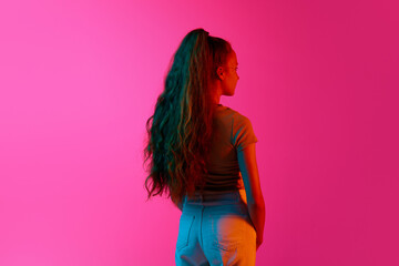 Back view of young girl with long curly hair in ponytail standing and looking away against pink...