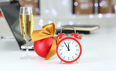 Red alarm clock and champagne goblet at office table in new year eve closeup. Festive mood within an inch of 2024 garland in background concept