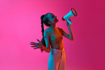 Young girl, student speaking in megaphone against pink studio background in neon light. News,...