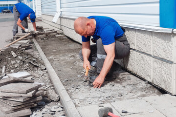 Elderly bricklayer lays tiles on foundation of building outside. Construction contractor lays out facing tiles of facade of production facility. Man lays tiles on summer day.