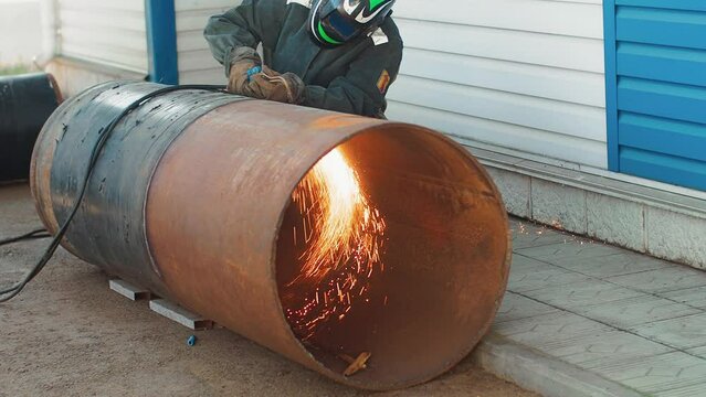 Welder in protective mask cuts large diameter pipe with autogenous gun from outside and sparks fly. Video of authentic production workflow.