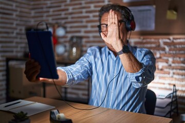 Middle age hispanic man using touchpad sitting on the table at night waiving saying hello happy and smiling, friendly welcome gesture