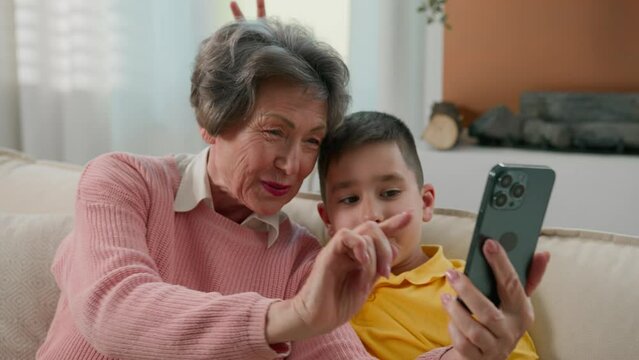 Caucasian grandmother and grandson together at home couch photographed selfie on mobile phone fool around funny family two generations grandma make vlog with kid boy taking pictures using smartphone