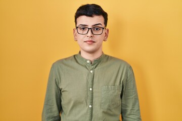 Non binary person standing over yellow background relaxed with serious expression on face. simple and natural looking at the camera.