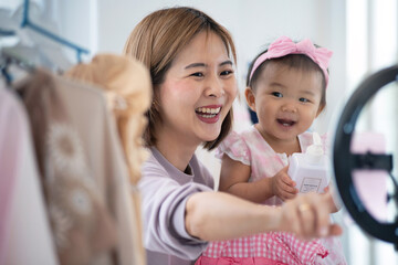 Obraz premium Asian female blogger live streams and shows her daughter's clothes and outfits to customers in front of a smartphone, online retail to sales at home, SME small online retail business at home