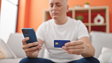 Young caucasian man shopping with smartphone and credit card sitting on sofa at home