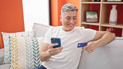 Young caucasian man shopping with smartphone and credit card sitting on sofa at home