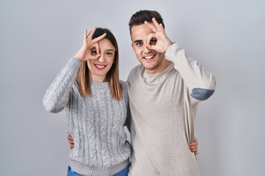 Young hispanic couple standing over white background doing ok gesture with hand smiling, eye looking through fingers with happy face.