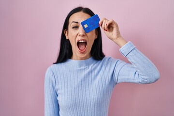 Hispanic woman holding credit card over eye angry and mad screaming frustrated and furious, shouting with anger. rage and aggressive concept.
