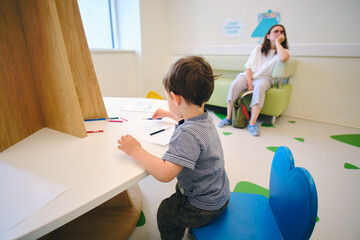 The child with her mother is patiently sitting in the hall of the medical center, drawing on a...