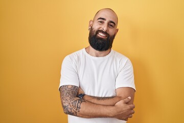 Young hispanic man with beard and tattoos standing over yellow background happy face smiling with...