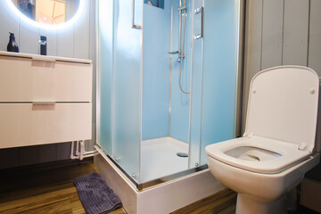 There's a contemporary shower cabin and a compact toilet bowl in the minimalistic bathroom. Round illuminated mirror over a white sink in a small bathroom
