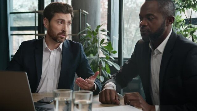 Two diverse multiracial businessmen in office work with computer data discuss startup online project with laptop. Caucasian businessman consulting African American man client, colleagues men together