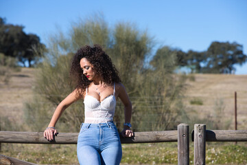 curly-haired, brunette, Spanish woman resting on the fence of the path leading to the meadow. She is dressed in jeans and a white top and enjoys the sunny day. The woman is sad and depressed.