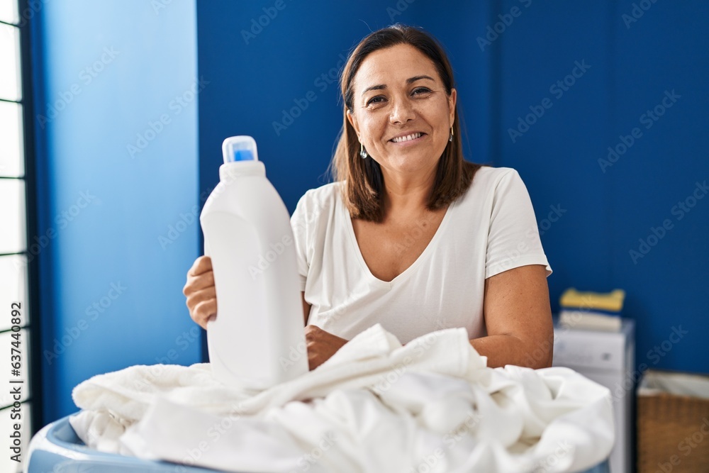 Wall mural Middle age hispanic woman doing laundry holding detergent bottle looking positive and happy standing and smiling with a confident smile showing teeth