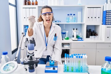 Young hispanic woman working at scientist laboratory angry and mad raising fist frustrated and furious while shouting with anger. rage and aggressive concept.