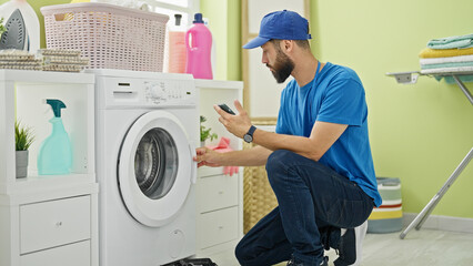 Young hispanic man technician repairing washing machine holding smartphone at laundry room