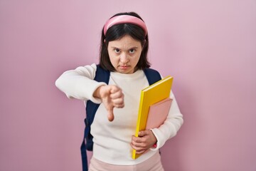 Woman with down syndrome wearing student backpack and holding books looking unhappy and angry showing rejection and negative with thumbs down gesture. bad expression.