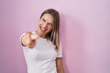 Blonde caucasian woman standing over pink background pointing displeased and frustrated to the...