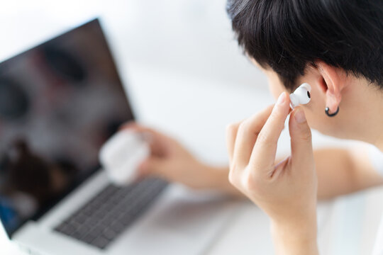 Asian man working on a personal laptop or notebook computer at public working space and using a truly wireless headphones or earbuds. Man using wireless earphones for listening music and conversation.