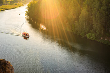 the boat floats on the river on a summer evening in the rays of the sun. forest along the banks of the river - obrazy, fototapety, plakaty