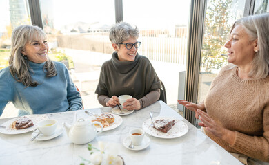 Three elderly women are sitting at a table in a coffee shop, talking and enjoying their tea.
