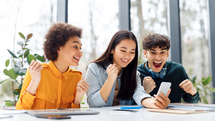 Obraz premium Excited multiracial students looking at cellphone and clenching fists, checking exams results online in classroom