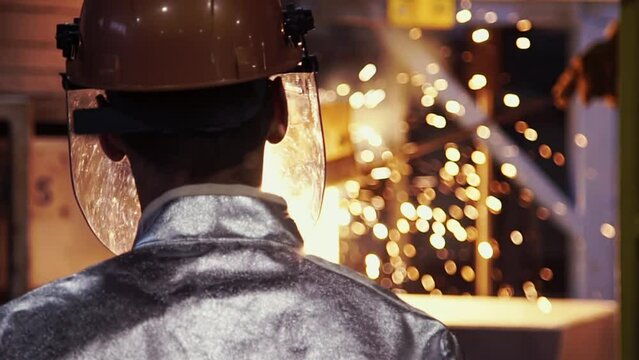 Cinematic rear view of factory worker in helmet at modern metal production plant. Foundry, metal cast, bright yellow sparks in the background, selective focus, bokeh, light. slow motion