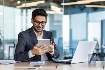Young successful hispanic businessman at workplace using app on tablet computer, smiling man inside office building, working with laptop, wearing business suit, happy with achievement. - obrazy, fototapety, plakaty