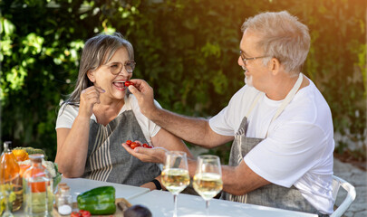 Happy old caucasian husband feeding his wife in apron with tomatoes at table with glasses of wine