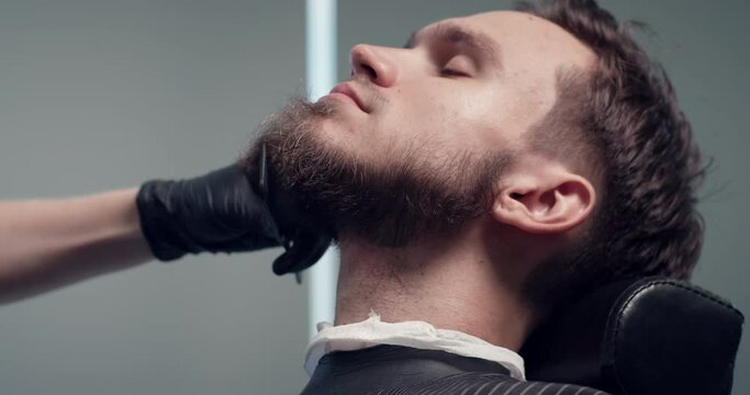 Barber trimming bearded man with shaving machine in barbershop. Hairstyling process. Close-up of a Hairstylist cutting the beard of a bearded male. 4K