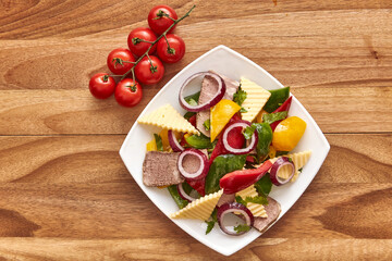White plate with salad of meat, cheese, bell pepper, red onion and herbs on the wooden table with cherry tomatoes near it. Top view, flat lay